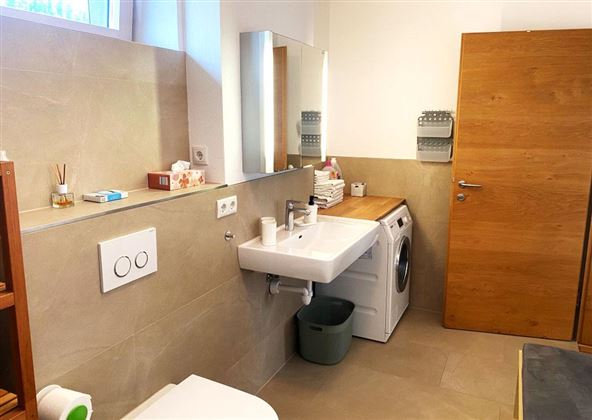 Een moderne badkamer met een wasmachine, een wastafel en houten planken. De muren zijn licht en de vloer is in neutrale tinten gehouden.