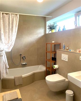 Een modern badkamer met een bad en een douche. Er is een houten plank en grote ramen die natuurlijk licht binnenlaten.