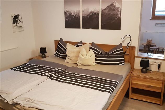 A cozy bedroom with a large bed and a minimalist design.  
On the wall hang pictures of mountains, and there are two nightstands with lamps.