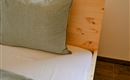 Een gezellig bed met een houten frame en een groot kussen. De beddengoed is in zachte kleuren uitgevoerd.