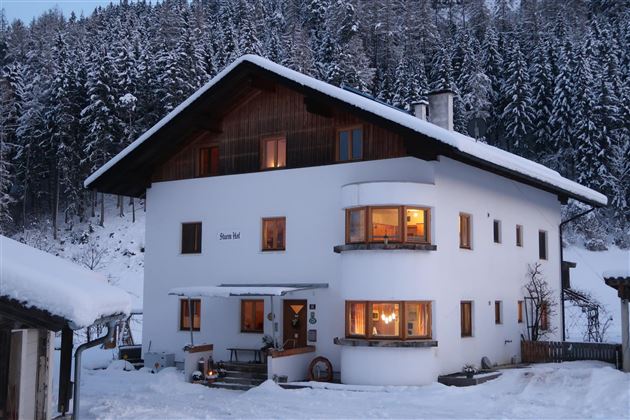 Una casa accogliente nella neve, circondata da alte montagne boscose. Le finestre brillano calde in contrasto con il freddo paesaggio invernale.
