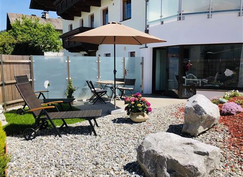 A cozy garden with a sunny seating area. A large beach umbrella provides shade, surrounded by flowers and stones.