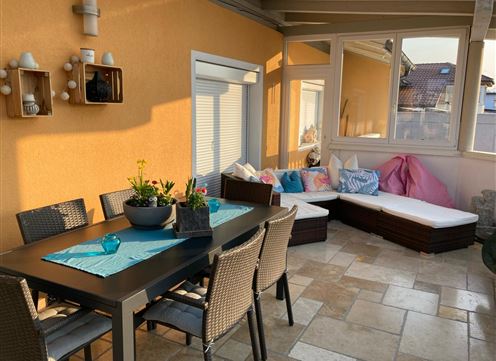 A cozy terrace with a dining area and a sofa corner. The walls are painted orange and there are plants as well as decorative cushions.