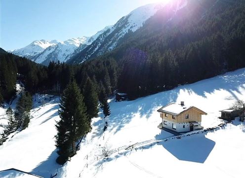A beautiful chalet in a snowy landscape, surrounded by tall firs and mountains. The sun shines over the snow-covered surroundings.