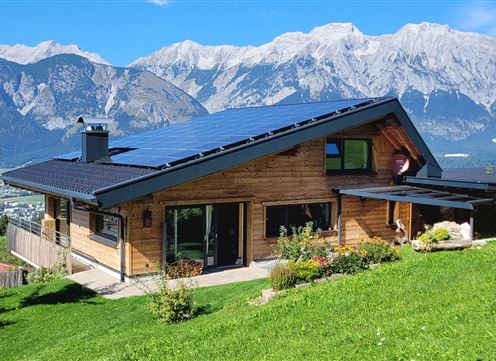 Ein modernes Holzhaus mit Solarpanelen in einer grünen Landschaft. Im Hintergrund erheben sich majestätische Berge unter einem klaren blauen Himmel.