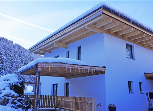 A modern house with a white facade and wood details. Winter has covered the surroundings with snow, and in the background, snow-covered trees are visible.