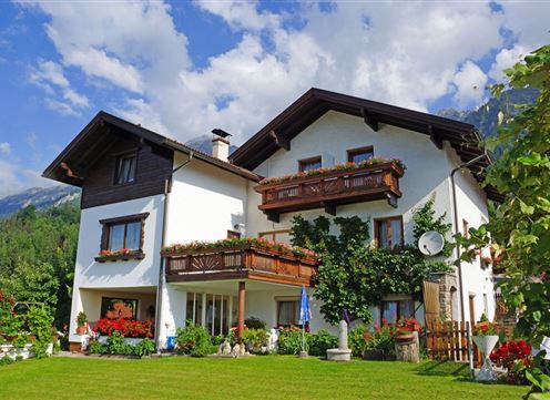 A beautiful two-story house with a balcony and many flowers. The garden is green and well-kept, surrounded by trees and mountains.
