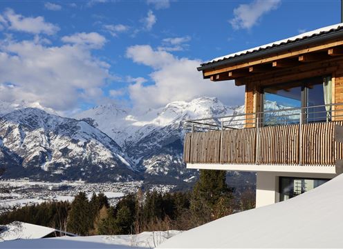 A modern mountain cabin with a balcony overlooks snow-covered mountains. The sky is blue with some clouds.