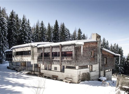 A modern wooden house in the snow, surrounded by tall, green fir trees. The architecture combines rustic charm with contemporary design.