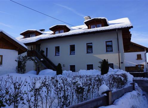 A cozy house in the snow with a clear blue sky. The surroundings are adorned with white snow and green shrubs.