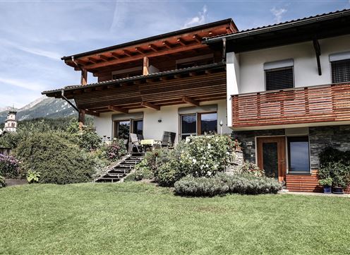 A beautiful house with a well-maintained garden and an inviting terrace. In the background, mountains and a blue sky can be seen.