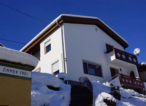 A modern house in the snow with a clear blue sky. The sign reads "Rooms available."