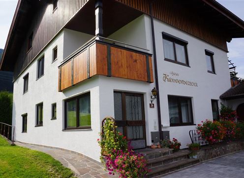 Ein charmantes weißes Ferienhaus mit einem Holzbalcony. Blumen schmücken den Eingangsbereich und die Umgebung ist grün und einladend.