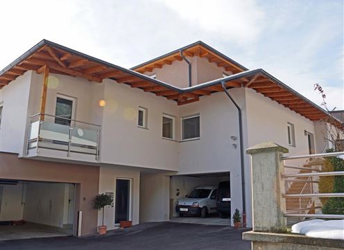 A modern two-storey house with a wooden overhang. In front of the building, there are some plants and a car is in the garage.