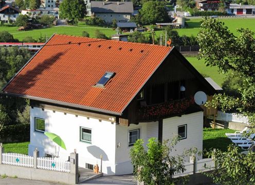 A beautiful house with a red roof in a green environment. There is a garden with loungers and a white fence.