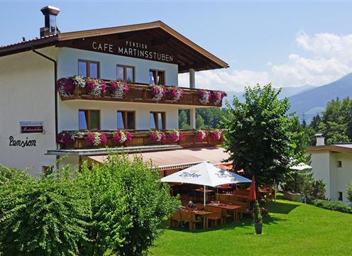 A cozy guesthouse with a balcony and blooming plants. In the foreground, there are outdoor seating areas surrounded by greenery.