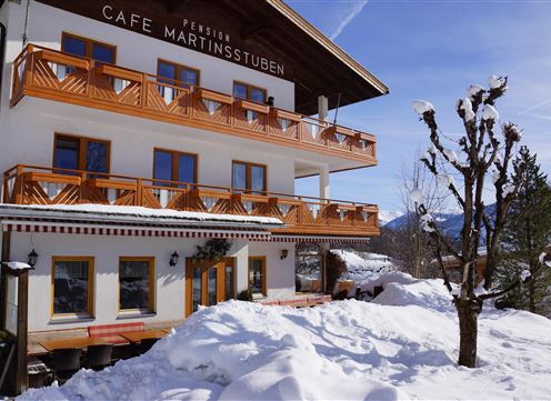 A cozy building in the snow with a sign that says "Café Martinstub'n". The surroundings are wintry with lots of snow and a clear sky in the background.
