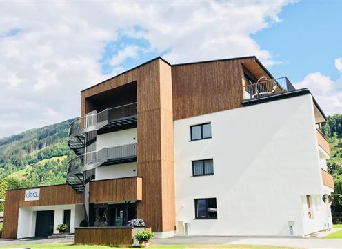 A modern building with wooden cladding and double balconies. It is located in a green, mountainous area.
