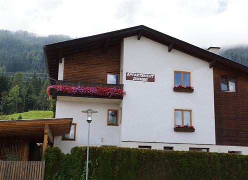Ein gemütliches Appartementhaus mit einem Holz- und Weißanstrich. Blumenkästen schmücken die Fenster, umgeben von malerischer Natur.