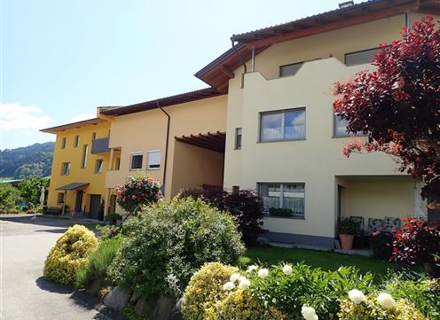 A modern building with a yellow facade and large windows. In front of the house, there are well-maintained garden facilities with colorful flowers.