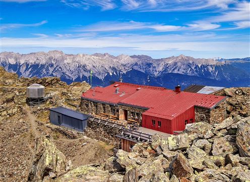 A mountain hut with a red roof on a rocky peak. In the background, majestic mountains and a blue sky can be seen.