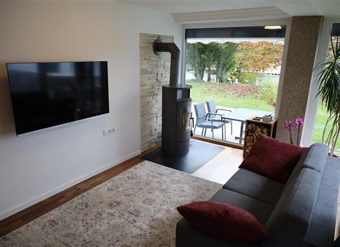 A modern living room with a gray sofa and a television on the wall.  
Through large windows, the view of the garden is visible.