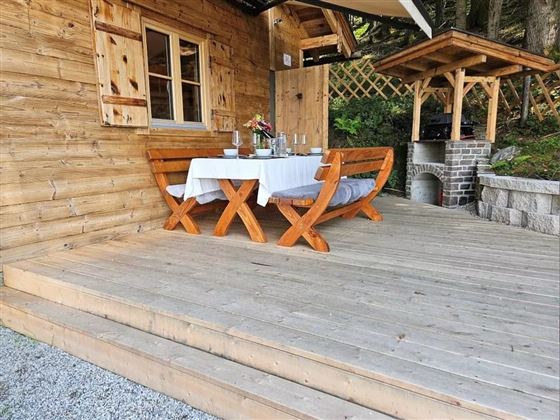 Eine gemütliche Terrasse aus Holz mit einem Tisch und Stühlen. Auf dem Tisch steht eine Blumenvase und es sind Gläser für Getränke bereitgestellt.
