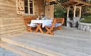 Eine gemütliche Terrasse aus Holz mit einem Tisch und Stühlen. Auf dem Tisch steht eine Blumenvase und es sind Gläser für Getränke bereitgestellt.