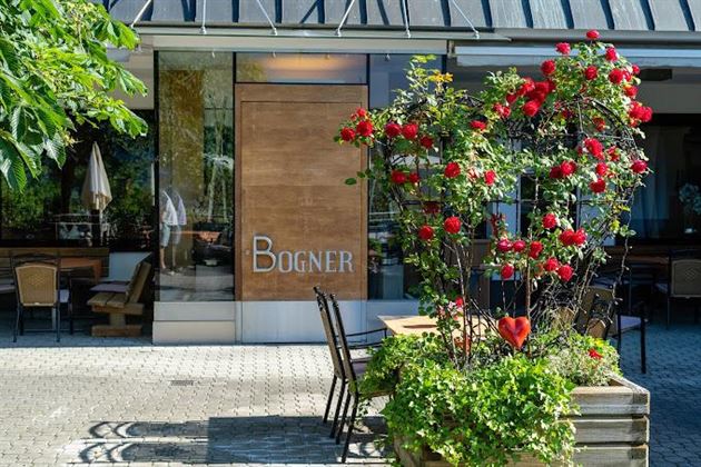 Un restaurant élégant avec l'inscription "Bogner". Devant l'entrée, des roses rouges fleurissent et il y a quelques tables en plein air.