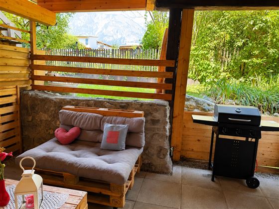 Eine gemütliche Sitzecke aus Paletten mit Kissen in einem einladenden Garten. Neben der Sitzecke steht ein Grill und im Hintergrund sind grüne Pflanzen und Bäume zu sehen.