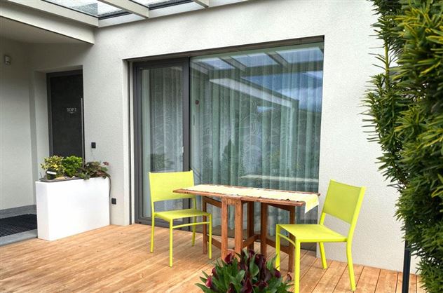 Een gezellige terras met een houten tafel en twee lichtgroene stoelen. Op de achtergrond zijn planten en grote ramen te zien.