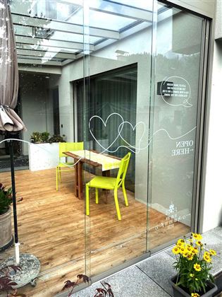 Een modern café met een houten vloer en grote glazen ramen. Binnen staan een tafel en twee lichtgroene stoelen, omringd door planten en bloemen.