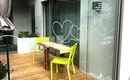 Een modern café met een houten vloer en grote glazen ramen. Binnen staan een tafel en twee lichtgroene stoelen, omringd door planten en bloemen.
