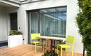 Een gezellige terras met een houten tafel en twee lichtgroene stoelen. Op de achtergrond zijn planten en grote ramen te zien.