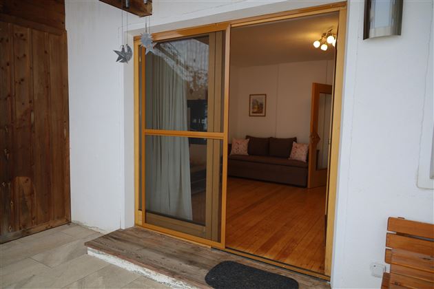 Ein einladender Eingangsbereich mit einer offenen Terrassentür. Im Hintergrund ist ein gemütliches Wohnzimmer mit einem Sofa und sanften Farben sichtbar.