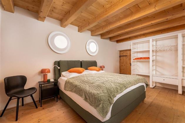 Ein modernes Schlafzimmer mit einem Doppelbett und zwei Kissen in Orange. Der Raum hat Holzbalken und große runde Fenster, sowie einen minimalistisch gestalteten Kleiderschrank.