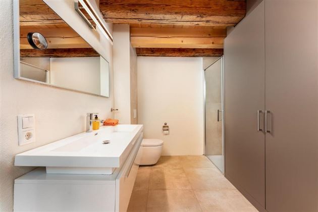 Ein modernes Badezimmer mit einer weißen Waschbeckenzeile und einem großen Spiegel. Die Wände sind hell und die Decke hat sichtbare Holzbalken.