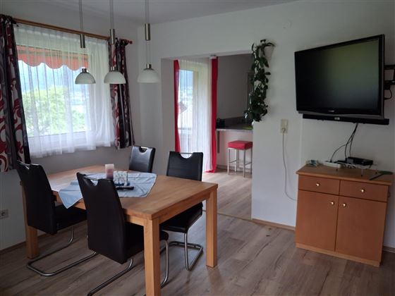 Een uitnodigende eetruimte met een houten bureau en zwarte stoelen. Aan de muur hangt een tv, en door het raam valt er veel daglicht naar binnen.