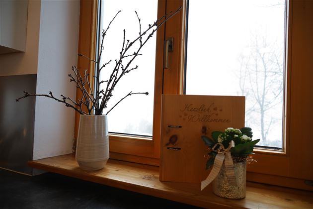 Ein Fenster mit einem breiten Fensterbrett. Darauf stehen eine Vase mit Zweigen und eine dekorative Box neben einem kleinen Blumengebinde.