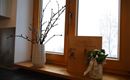 Ein Fenster mit einem breiten Fensterbrett. Darauf stehen eine Vase mit Zweigen und eine dekorative Box neben einem kleinen Blumengebinde.