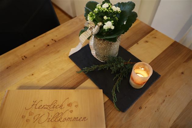 Ein herzlich dekorierter Tisch mit einer Pflanze in einer Glaskaraffe, einem Teelicht und einem Willkommensschild. Die warmen Holz- und Grüntöne schaffen eine einladende Atmosphäre.