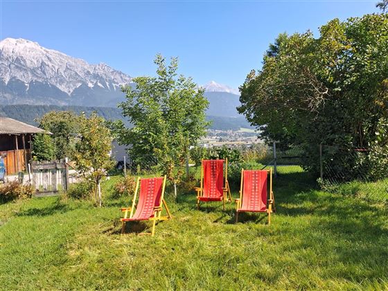 Een zonnige tuin met drie rode ligstoelen en uitzicht op de bergen. De natuur is groen en uitnodigend.