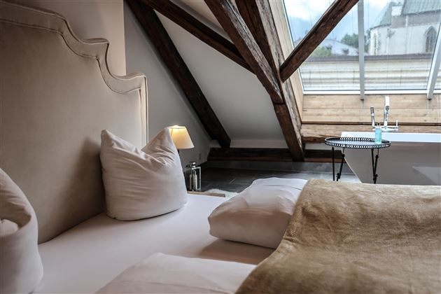 Une chambre confortable avec des poutres en bois inclinées et une grande fenêtre. Le mobilier est moderne et accueillant.