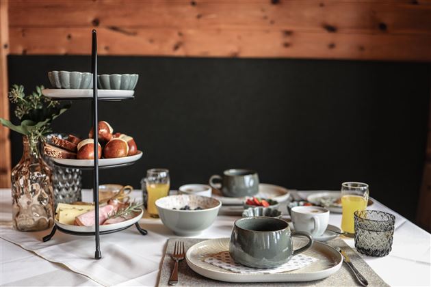 Eine schön gedeckte Frühstückstafel mit verschiedenen Speisen und Getränken. Auf einem dreistöckigen Servierständer sind Brötchen und andere Leckereien angerichtet.