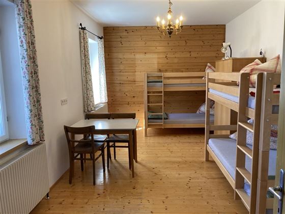 A simple room with a bunk bed and a table with chairs. The walls are light, and there are large windows with curtains.