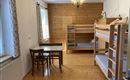A simple room with a bunk bed and a table with chairs. The walls are light, and there are large windows with curtains.