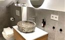 Une salle de bains moderne avec un élégant lavabo en pierre sur une commode en bois. La pièce est lumineuse et minimaliste avec des carreaux gris et un miroir rond.