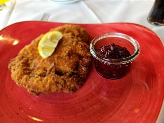 A golden-brown breaded schnitzel lies on a red plate, garnished with a slice of lemon. Next to it is a small glass of jam.