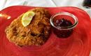 A golden-brown breaded schnitzel lies on a red plate, garnished with a slice of lemon. Next to it is a small glass of jam.