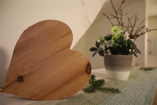Ein hölzernes Herz steht auf einem Tisch neben einem Topf mit grünen Pflanzen. Der Tisch ist mit einem gestreiften Tuch bedeckt.